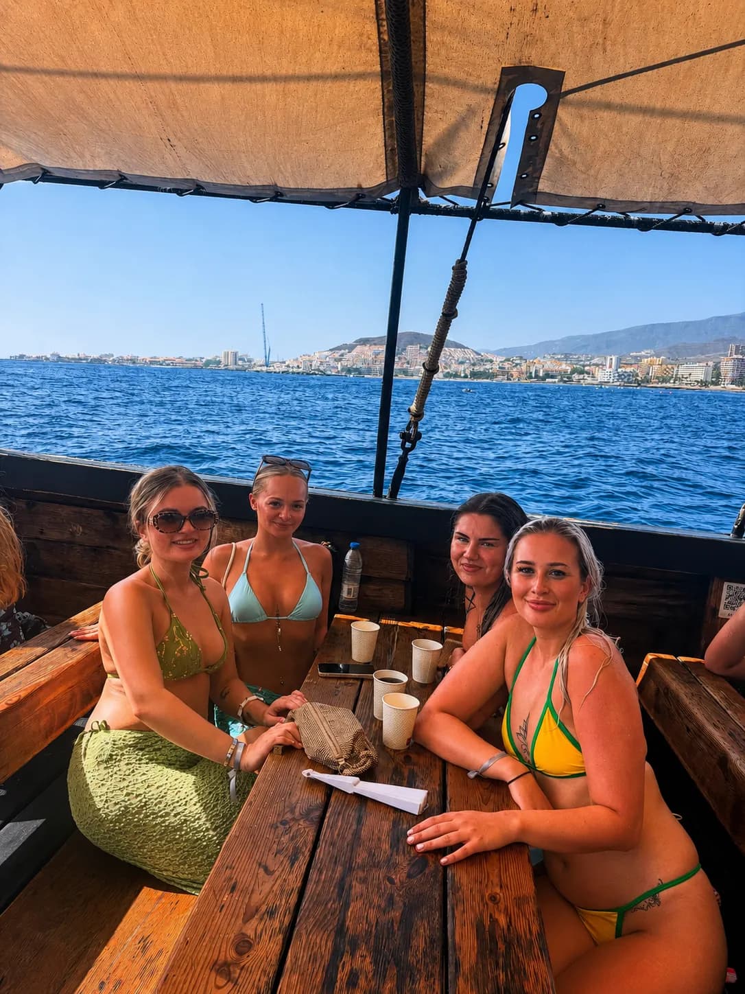 Four young women smiling on wooden Tenerife boat deck with coastal town backdrop