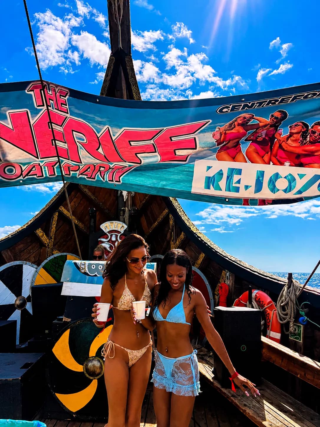 Two women in bikinis holding drinks on pirate-themed boat deck